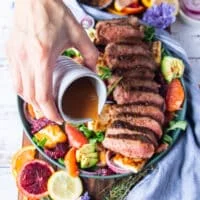 A hand drizzling the honey citrus dressing over the roast lamb loin and halloumi salad to finish the dish