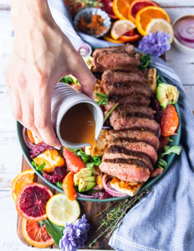 A hand drizzling the honey citrus dressing over the roast lamb loin and halloumi salad to finish the dish