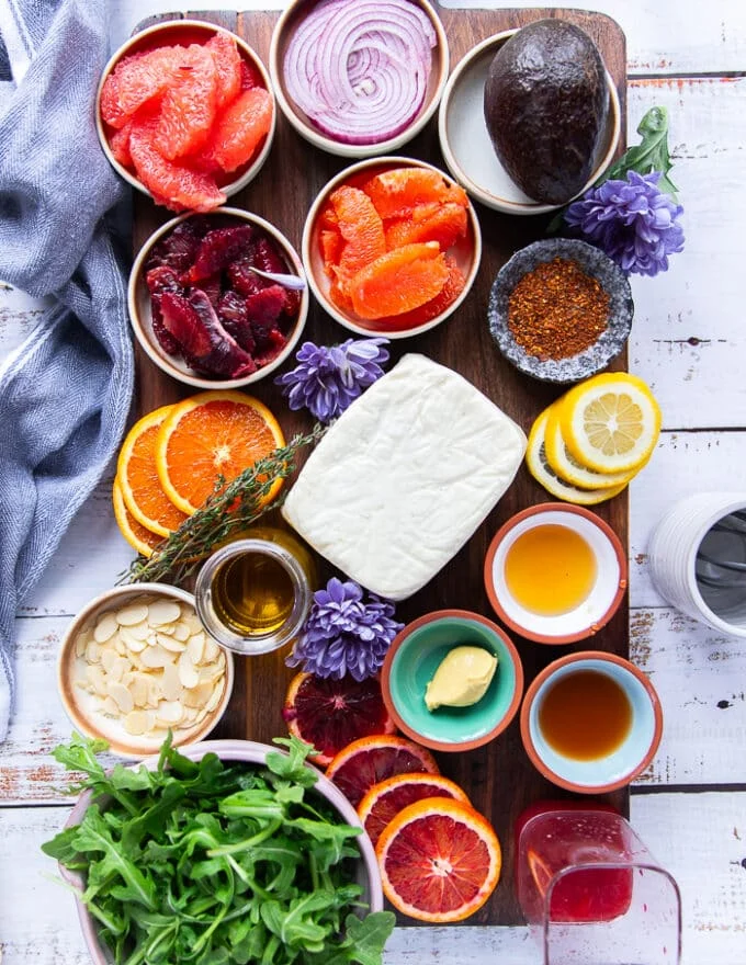 The halloumi salad ingredients and citrus dressing on a cutting board including a block of halloumi cheese, a bowl of segmented orange, segmented blood orange, segments of grapefruit, some almonds in a bowl, a mustard in a bowl, a bowl of honey, a bowl of apple cider vinegar, a jar of olive oil, arugula