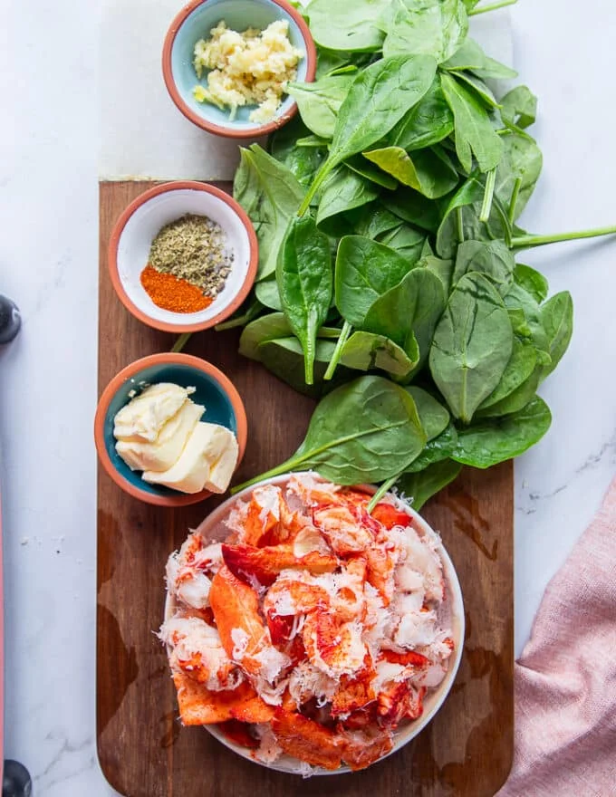 Ingredients for the lobster pasta on the cooking board including a bowl of cooked lobster tail, some garlic in a bowl, fresh cream, spinach in a bowl, sun dried tomatoes on a plate, unsalted butter in a plate and spices