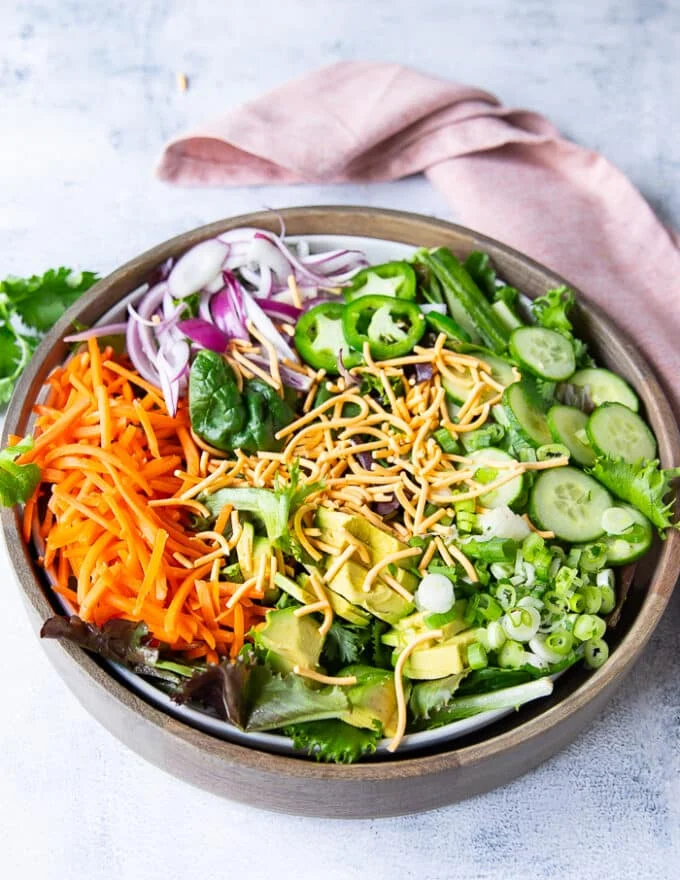 Salad ingredients including some cucumbers, some shredded carrots, greens, green onions, crispy wontons, red onions, avocados, cilantro, jalapenos