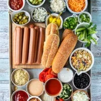 Hot Dog Bar ingredients including small bowls of many different tppinsg like cheese, veggies, sauces, a variety of pickles, breads, spices and more.
