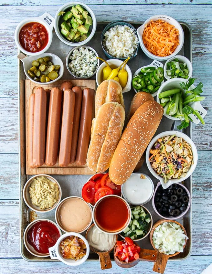 Hot Dog Bar ingredients including small bowls of many different tppinsg like cheese, veggies, sauces, a variety of pickles, breads, spices and more.