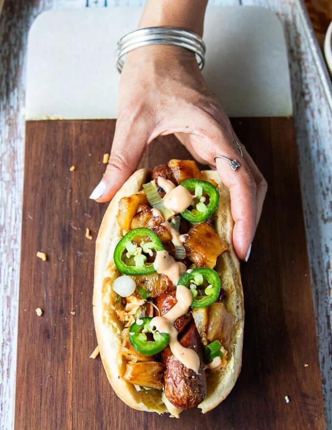An assembled Hawaiian style hot dog showing the grilled hot dog with spicy slaw, jalapenos and grilled pineapples