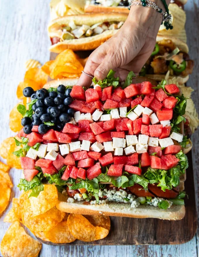 A hand holding a a board of watermelon salad shaped like the flag with feta and blueberries as an option to serve with grilled hot dogs and some chips on the board showing different options
