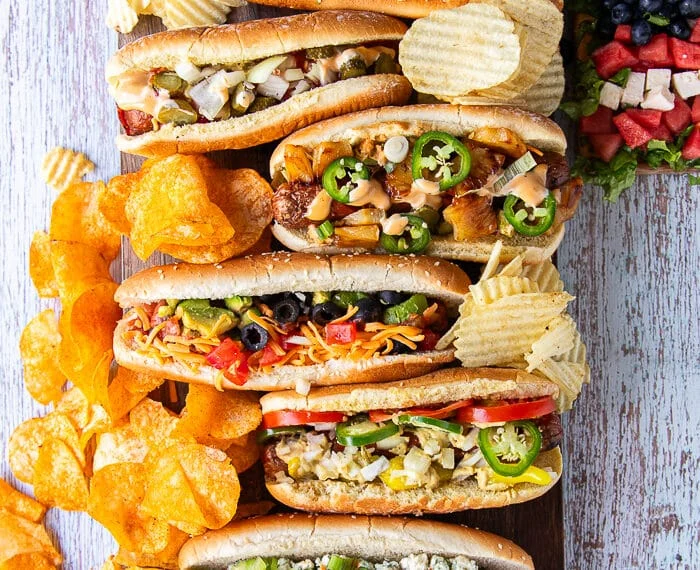 A board with 6 different assembled grilled dogs from the hot dog bar showing the variety of toppings. Some chips and a watermelon salad surrounding the board