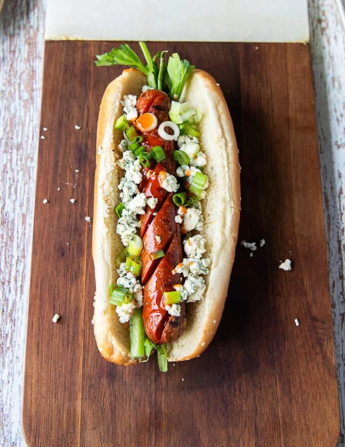 An assembled buffalo style hot dog showing the grilled hot dog with blue cheese, celery, hot sauce and green onions