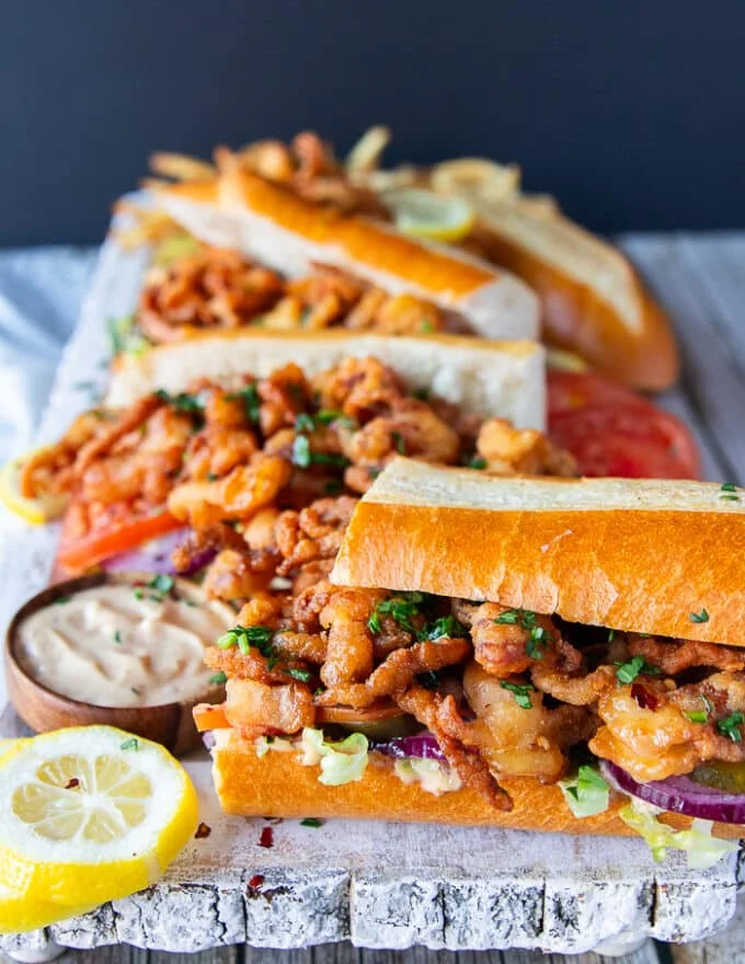 A board with a few Po Boy sandwiches surrounded by dipping sauce, lemon slices and close up of the fried shrimp showing the in front of the image