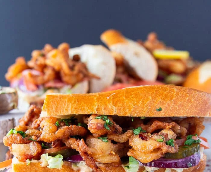 close up on a po boy sandwich cut in half , showing up close the fried shrimp and the sandwich toppings along with the sauce dripping on the sides of the bread