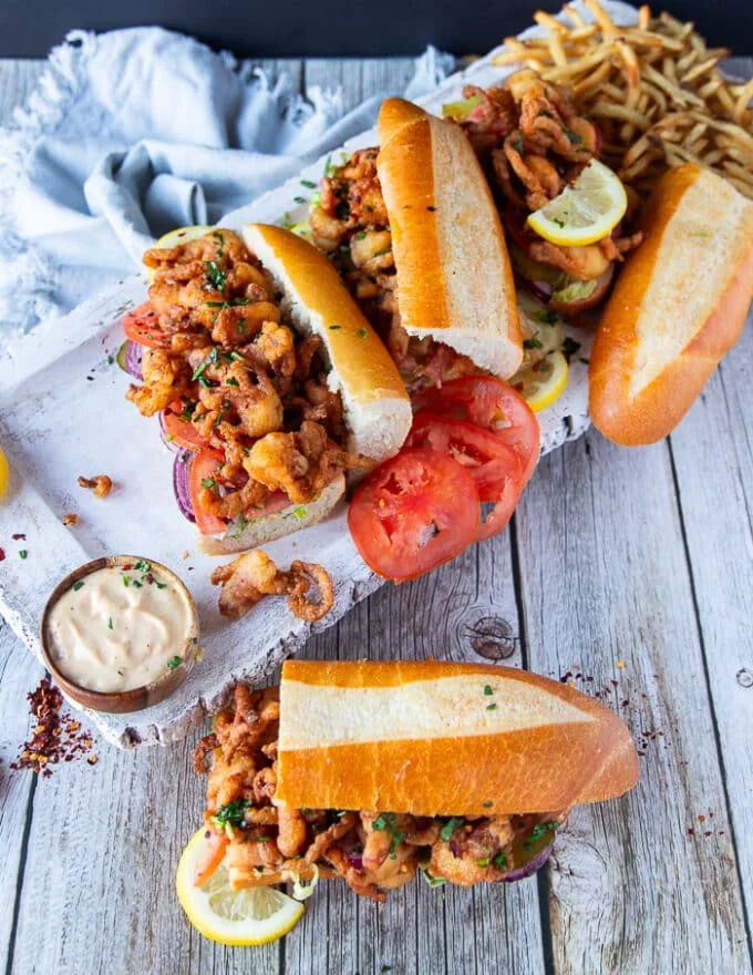 top view of Po Boy sandwiches scattered on a wooden board with dipping sauces, some fries, lemon slices, and french fries showing a feast of sandwiches for a crowd