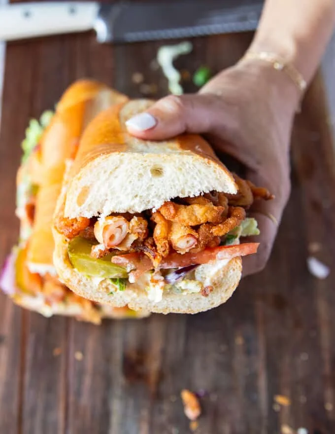 Po Boy Sandwich cut in half showing close u the texture and layers of sauce, lettuce tomatoes, pickles, onions and fried shrimp