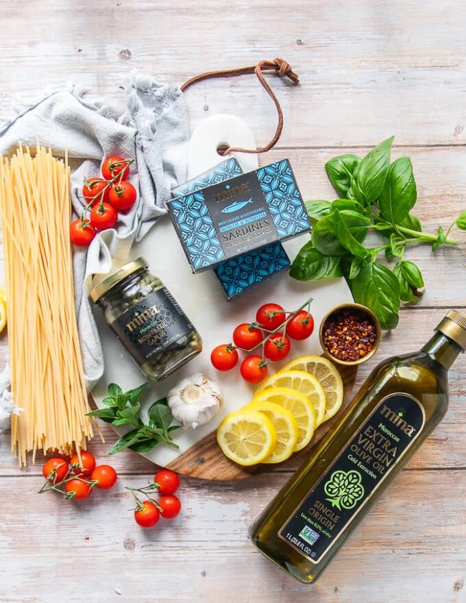Ingredients for sardine pasta including canned sardines, fresh lemon, lots of garlic, a bottle of olive oil, some capers, cherry tomatoes, some fresh basil, salt and pepper, and some uncooked pasta on the side