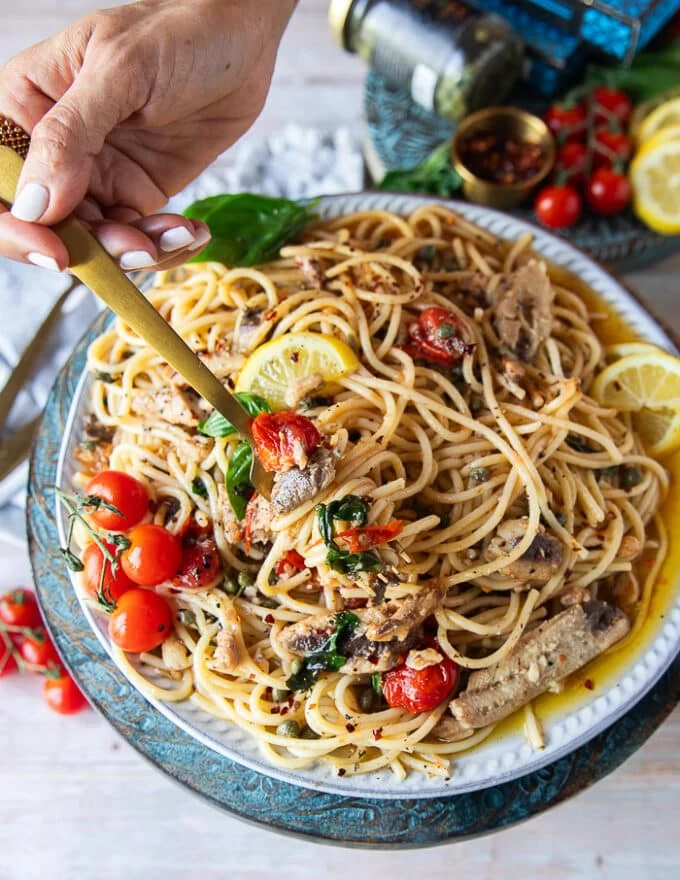 A hand holding a fork on a plate of sardine pasta showing the sardines, basil, tomatoes, capers, lemon and olive oil sauce around it