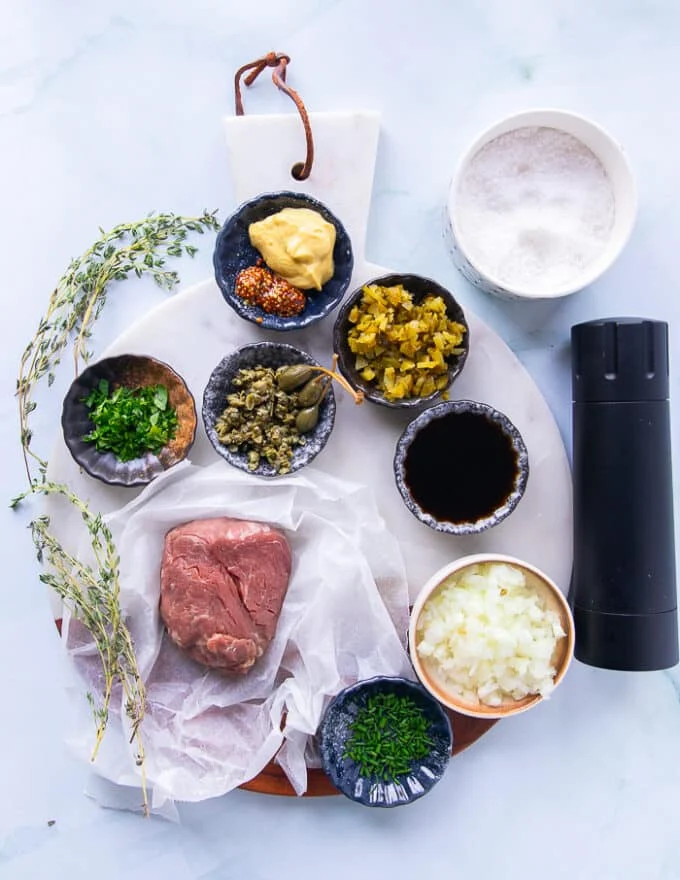 Ingredients for the steak tartare recipe on a white board including a piece of fresh beef tenderloin, a bowl of chopped onions, cowl of chopped capers, bowl of minced herbs, bowl of dijon and grainy mustard, bowl of Worcestershire sauce, salt and pepper and a bowl of chopped cornichons