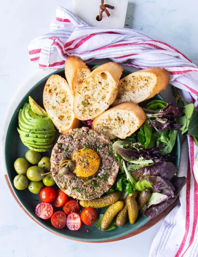 final plate of steak tartare with an egg yolk on top surrounded by some greens, baguette, cornichons, tomatoes, olives and avocados