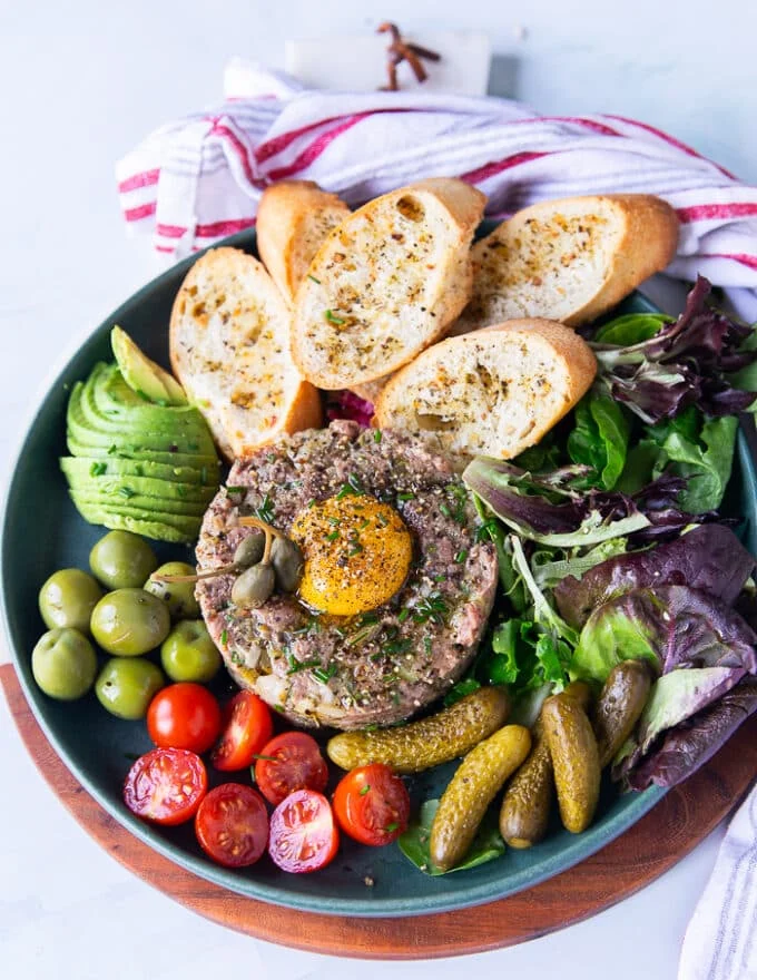 final plate of steak tartare with an egg yolk on top surrounded by some greens, baguette, cornichons, tomatoes, olives and avocados