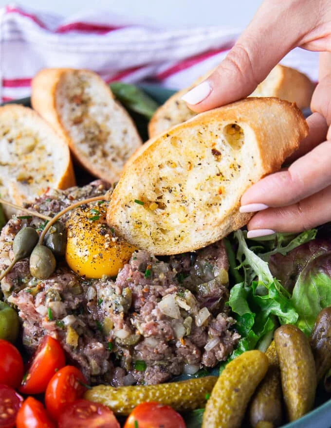 A hand holding a piece of crunchy baguette and pressing it on the eg yolk on stop of the steak tartare to break through and eat