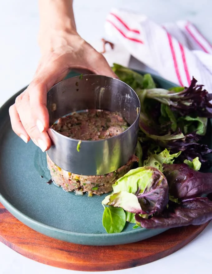A hand gently lifting the mold showing the beef tartare so beautifully stacked and round