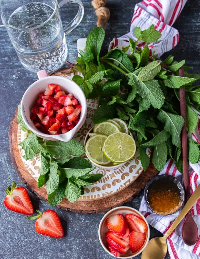 All ingredients you'll need to make strawberry mojito including strawberries, lime, lots of fresh mint, ice cubes, honey or simple syrup and soda water