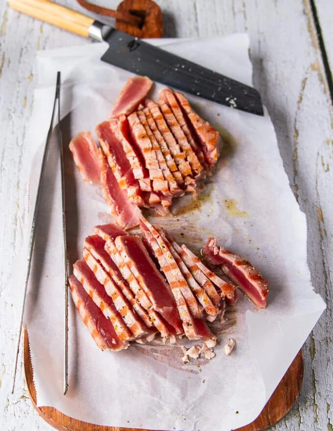 A sharp knife on a cutting board slicing the tuna super thin , showing how pink the tuna is on the inside and the beautiful crust