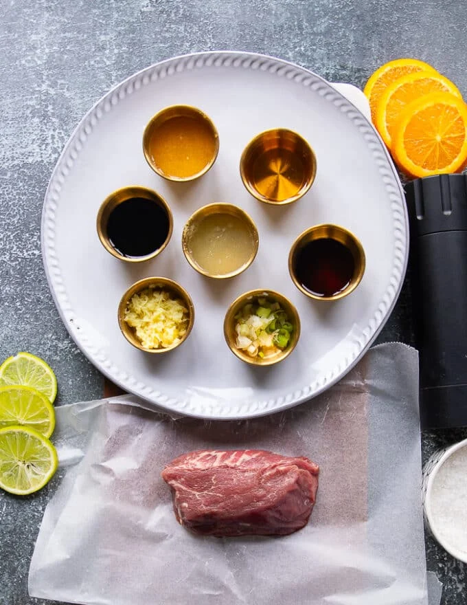 Ingredients needed to make beef tataki showing a piece of beef tenderloin, small bowls of ponzu ingredients like sesame oil orange juice, lime juice, soy sauce, brown sugar, grated ginger, Worcestershire sauce
