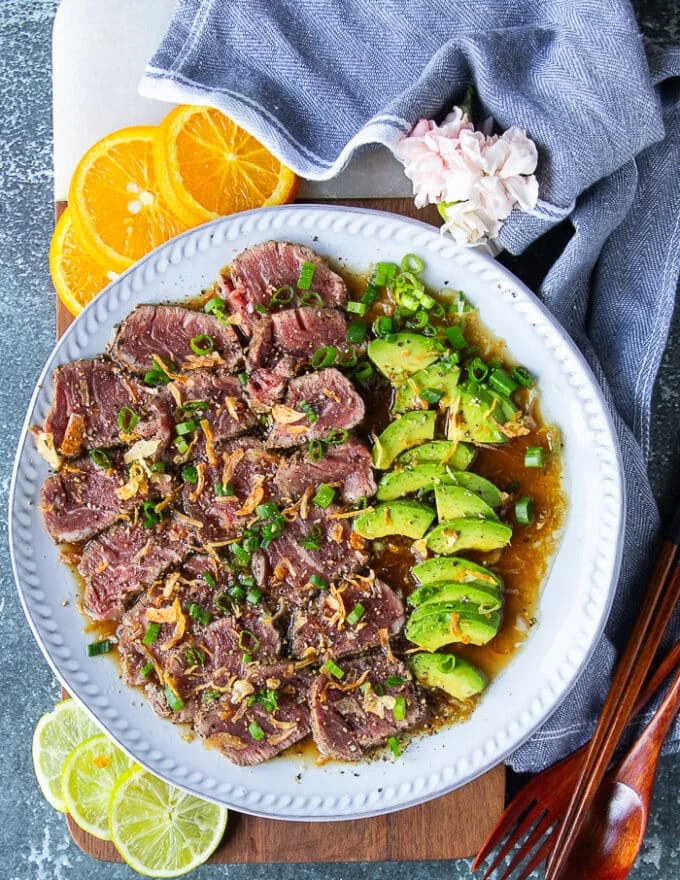 finished plate of beef tataki served over ponzu sauce with sliced avocados and garnished with fried onions and green onions