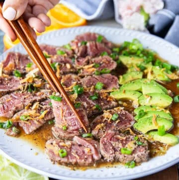 A hand holding two chopsticks and serving beef tataki sliced over a plate of ponzu sauce topped with crisp fried onions and green onions, surrounded by avocados sliced