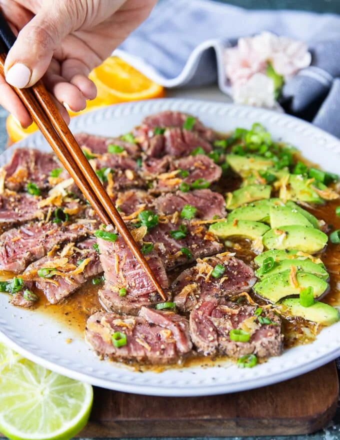 A hand holding two chopsticks and serving beef tataki sliced over a plate of ponzu sauce topped with crisp fried onions and green onions, surrounded by avocados sliced