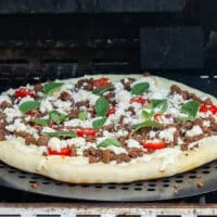 pizza on the grill with all the toppings ready like sausage, cheese, tomatoes and herbs