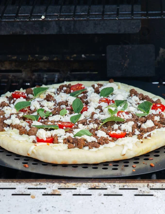 pizza on the grill with all the toppings ready like sausage, cheese, tomatoes and herbs