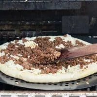 The pizza dough on the grill flipped and a hand adding toppings over the dough