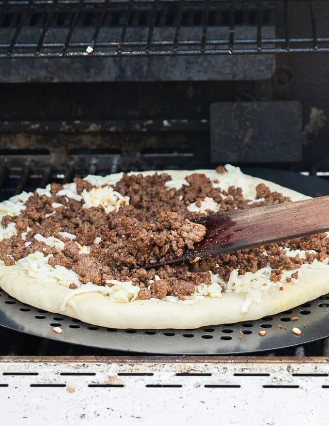 The pizza dough on the grill flipped and a hand adding toppings over the dough