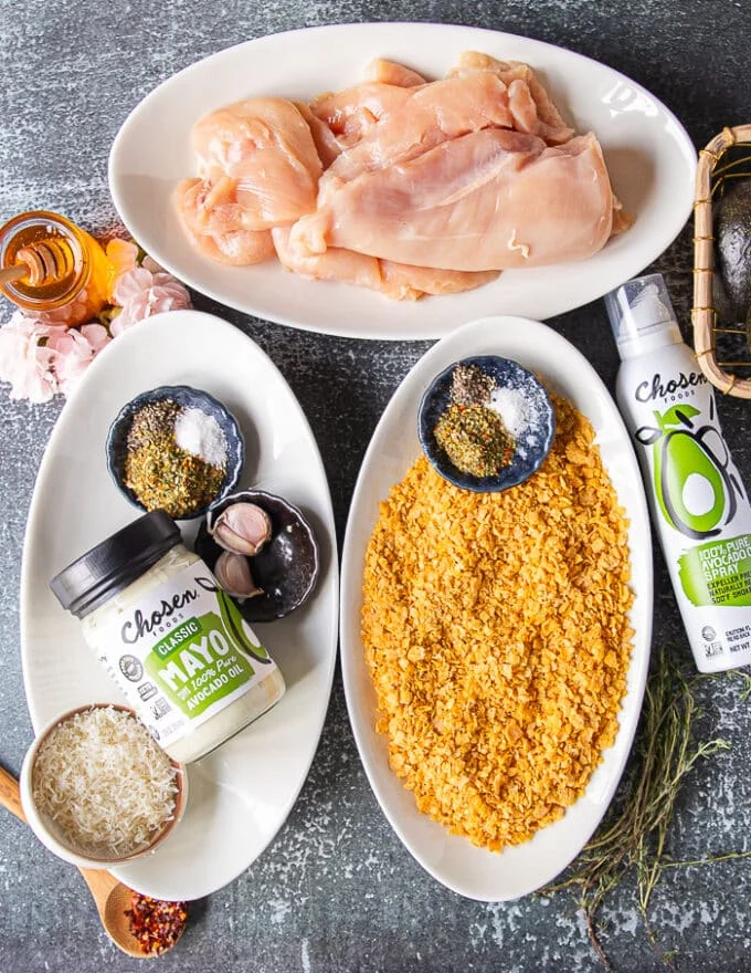 Ingredients for hot honey chicken tenders on a board including a bowls of chicken tenders, a bowl of corn flakes with a small bowl of spice, another plate with mayo, a bowl of spices, one egg, fresh garlic and some fresh parm