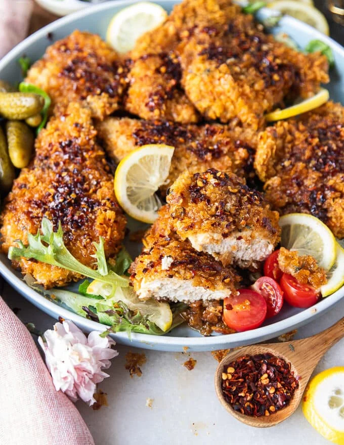 A plate with hot honey chicken tenders surrounded by lemon slices, a jar of hot honey and one chicken cut in half showing the juicy chicken inside.