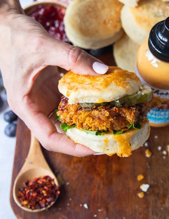 A hand holding a hot honey chicken sandwich showing the layers of hot honey chicken, chicken sauce, pickles, lettuce in a biscuit