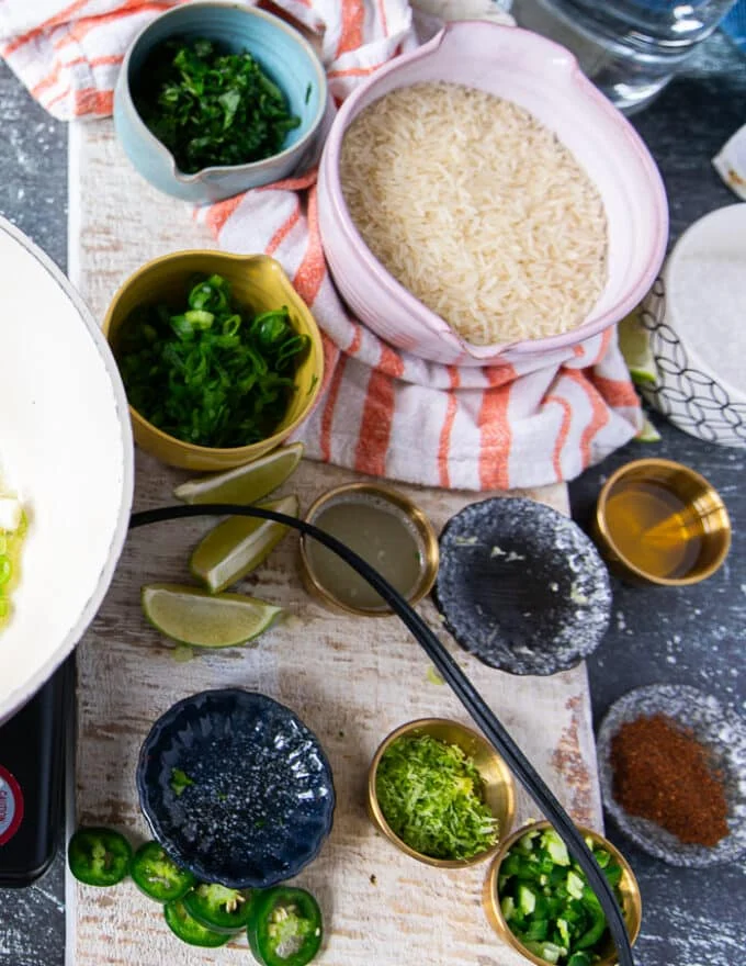 ingredients to make cilantro lime rice on a board including the bowl of rice, some olive oil in a bowl, some chopped cilantro, chopped scallions, lime zest, lime juice in bowls and chilli powder, some jalapeno minced