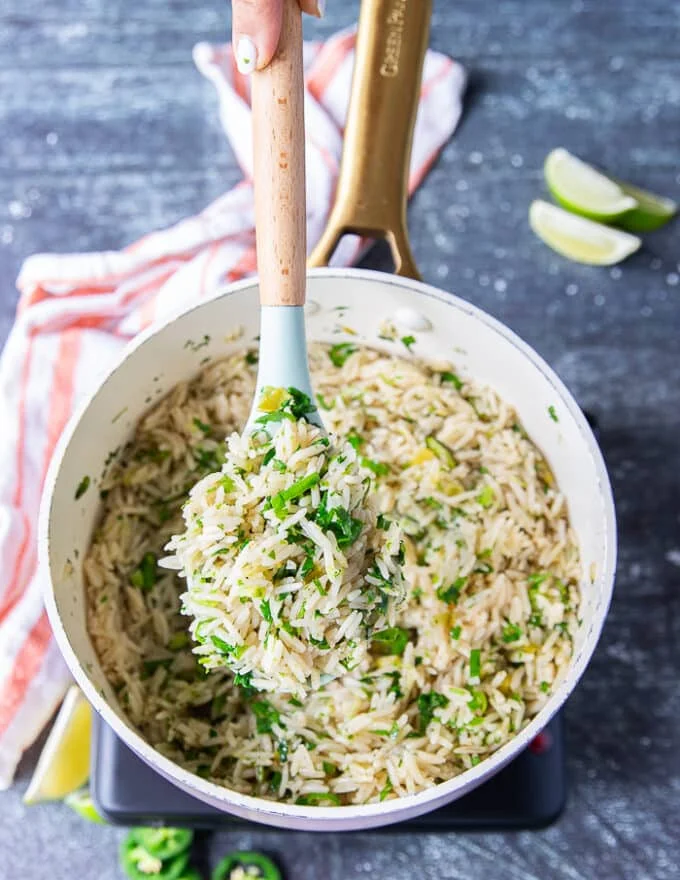 A spatula showing a spoonful of cilantro lime rice in the pot or rice cooker when tossed with the final lime zest, lime juice, cilantro , jalapeno and scallions showing specks of green and fluffy cooked white rice