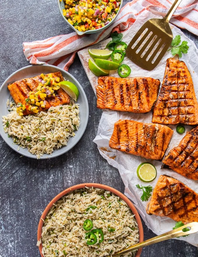 A set up of a plate full of grilled trout, a plate of cilantro lime rice to serve with, and a bowl of mango pico de gallo for a grilled fish feast
