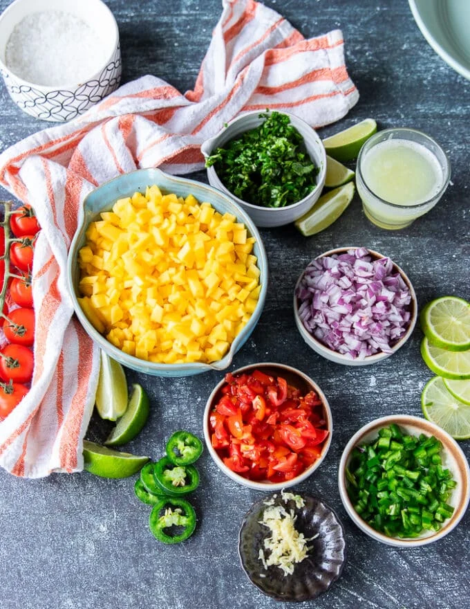Ingredients for mango pico de gallo including a bowl of chopped mangos, a owl of onions, cilantro leaves, some lime juice, a bowl of chopped tomatoes, bowl of jalapenos minced, some minced garlic and salt