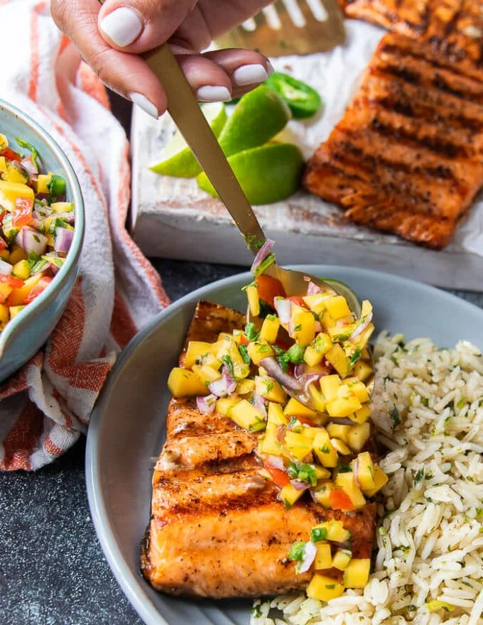 A hand spooning some mango pico de gallo over some grilled salmon fillets
