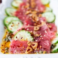 close up shot showing the tuna thinly sliced and sititng in the vinaigrette with a slice of cucumber pn the side, some garlic chips and capers around it. The texture of the tuna crudo is showing so clealry