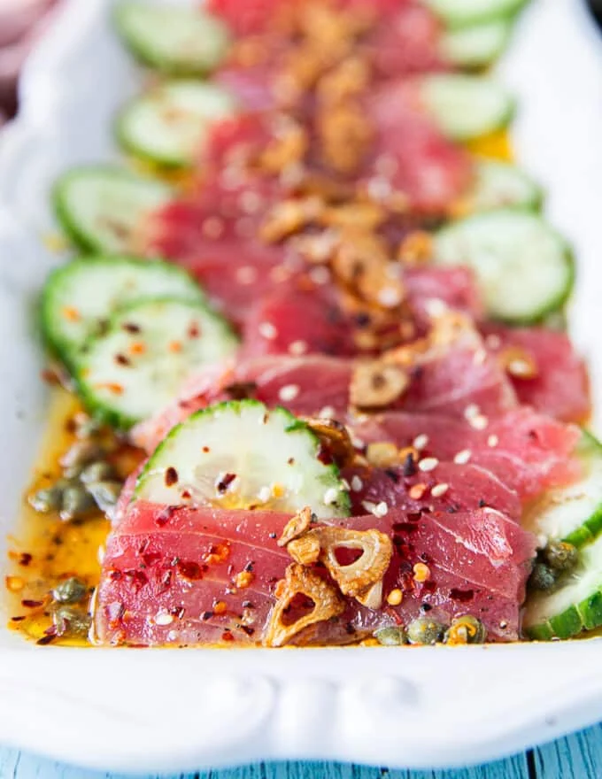 close up shot showing the tuna thinly sliced and sititng in the vinaigrette with a slice of cucumber pn the side, some garlic chips and capers around it. The texture of the tuna crudo is showing so clealry