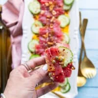 A hand holding a piece of tuna, cucumber over a crunchy Italian ciabbata bread showing how to serve tuna crudo with bread