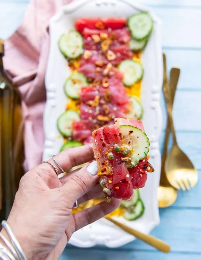 A hand holding a piece of tuna, cucumber over a crunchy Italian ciabbata bread showing how to serve tuna crudo with bread