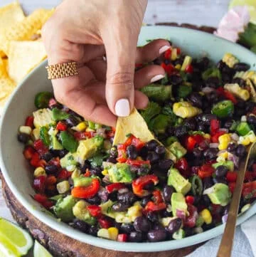 A hand holding a tortilla chip and scooping up some black bean and corn salad showing close up of the salad ingredients