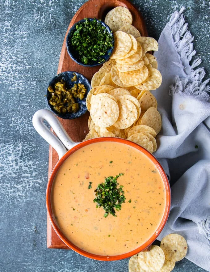 A bowl of queso on a board ready to be served along some tortilla chips, some extra jalapeno and cilanteo on the side optional for those who want to add it