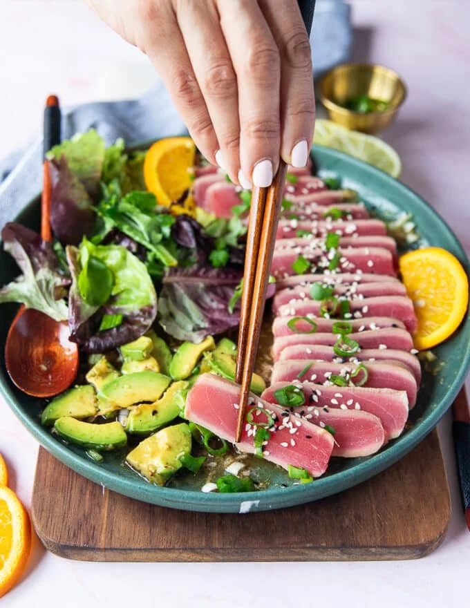 A hand holding one piece of tuna tataki with chopsticks up close showing the texture and ponzu dressing on top