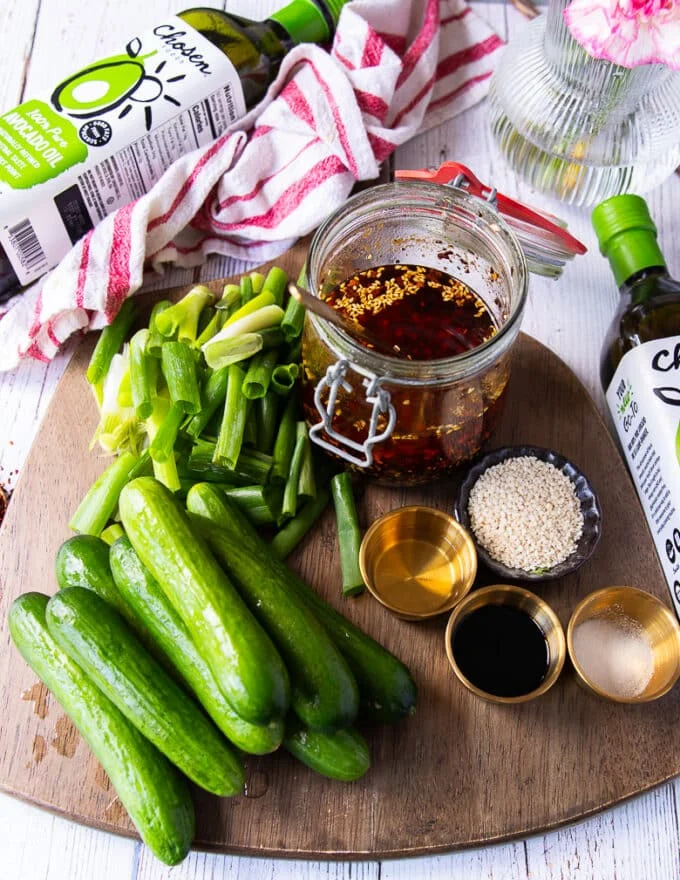 Asian cucumber salad ingredients in a cutting board including mini cucumbers, chili oil, sesame seeds, rice vinegar, soy sauce, garlic, scallions, ginger in different bowls