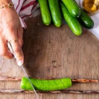 A hand holding a paring knife and making slits along the cucumber 1/4 inch apart on a 45 degree angle