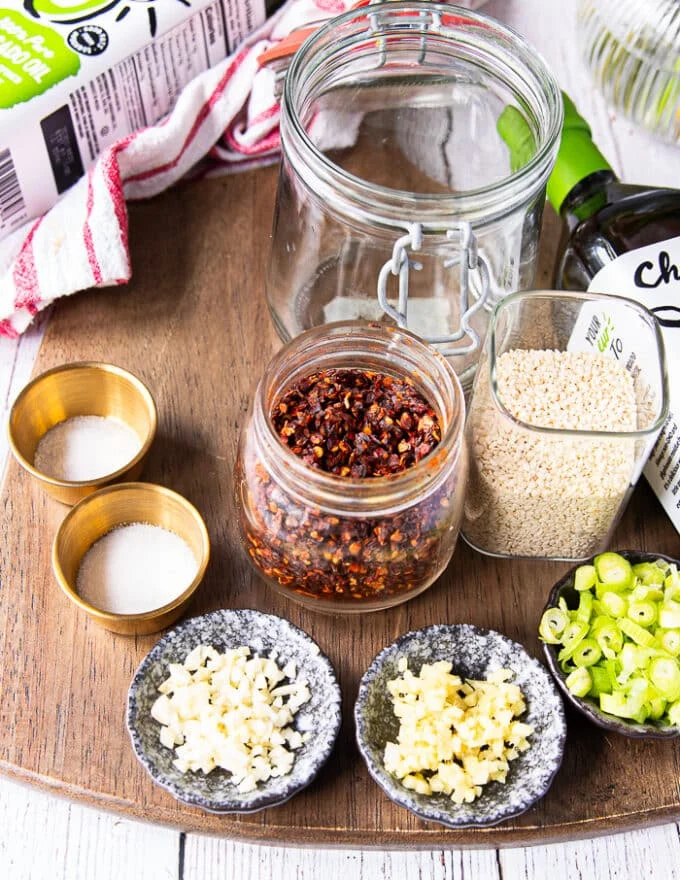 ingredients for chili oil recipe on a wooden board including a bowl of ginger, a bowl of garlic, a jar of sesame seeds, a jar of chilli flakes, a bowl of salt and one for sugar, some scallions minced, some oil